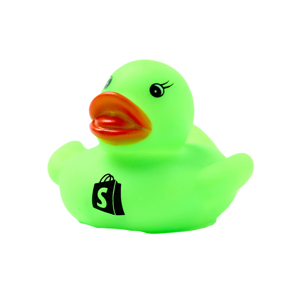 A close-up image of a Shopify Dev Duck, a bright green rubber duck with a small orange beak and cartoonish eyes. The duck features a black Shopify logo resembling a shopping bag on its side and is set against a white background.