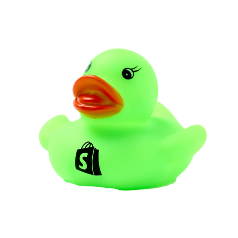 A close-up image of a Shopify Dev Duck, a bright green rubber duck with a small orange beak and cartoonish eyes. The duck features a black Shopify logo resembling a shopping bag on its side and is set against a white background.